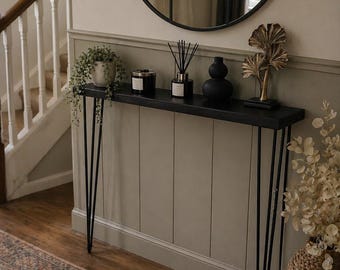 May include: A black console table with hairpin legs, adorned with decorative items. A round black mirror hangs above the table. The table holds a potted plant, candle, diffuser, black vase, and gold leaf sculpture. A patterned rug is on the floor.