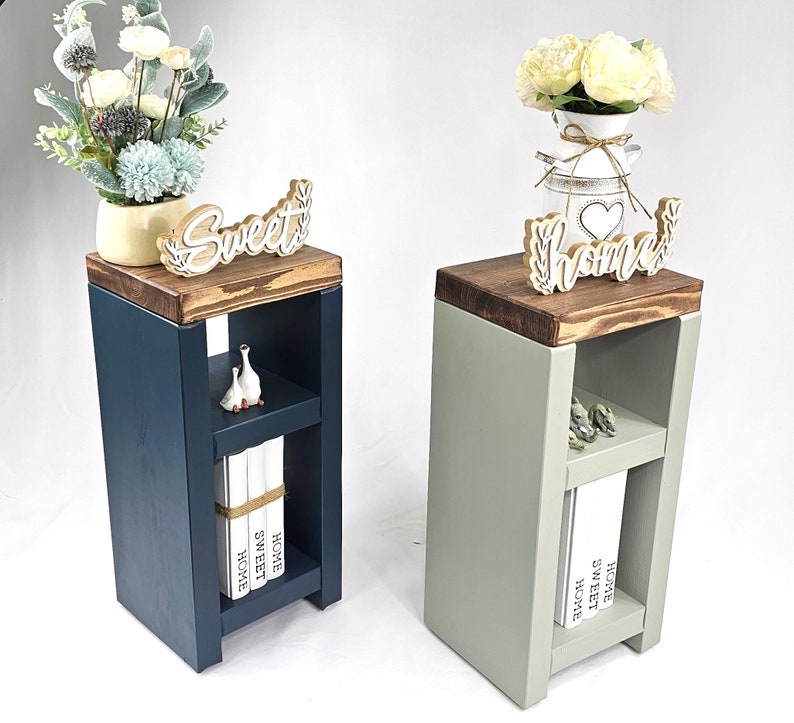 May include: Two wooden side tables, one navy blue and one sage green, each with a dark brown top. The navy table has a "Sweet" sign, while the green table has a "Home" sign. Both have a shelf with decorative items and books.