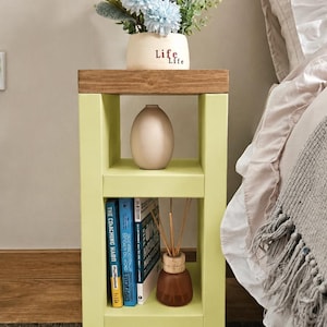 May include: A light green bedside table with a wooden top. It features a vase with blue and white flowers, a neutral-colored vase, books, and a small brown bottle with reeds. The table is on a patterned rug.