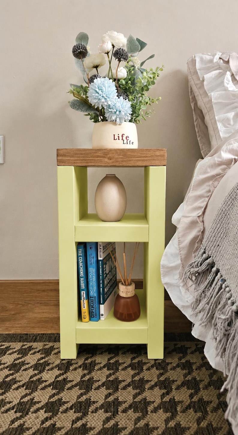 May include: A light green bedside table with a wooden top. It features a vase with blue and white flowers, a neutral-colored vase, books, and a small brown bottle with reeds. The table is on a patterned rug.