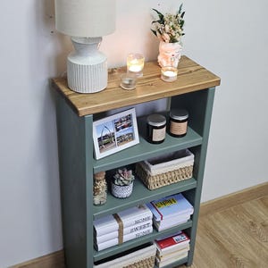 A wooden painted bookshelf with candles, framed photos, two baskets, decorative jars, and stacked books  placed on a wooden floor against a white wall.