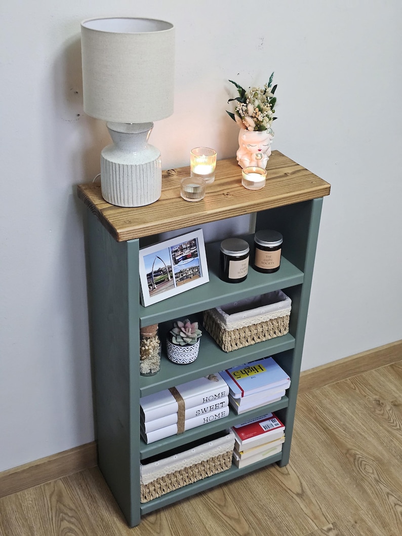 A wooden painted bookshelf with candles, framed photos, two baskets, decorative jars, and stacked books  placed on a wooden floor against a white wall.