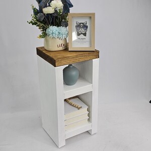 May include: A white wooden side table with a dark brown top. The table has a shelf with a stack of books and a small blue vase. A framed picture of a cat wearing sunglasses and a vase of flowers sit on top.