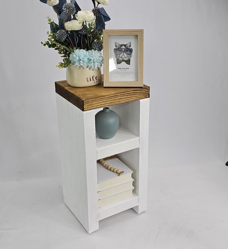 May include: A white wooden side table with a dark brown top. The table has a shelf with a stack of books and a small blue vase. A framed picture of a cat wearing sunglasses and a vase of flowers sit on top.