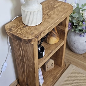May include: A small, rustic wooden side table with two shelves. A white lamp with a beige lampshade sits on top. The shelves hold a wooden sculpture, a black diffuser, a miniature house, and a small stack of cards. The table is made of light brown wood.