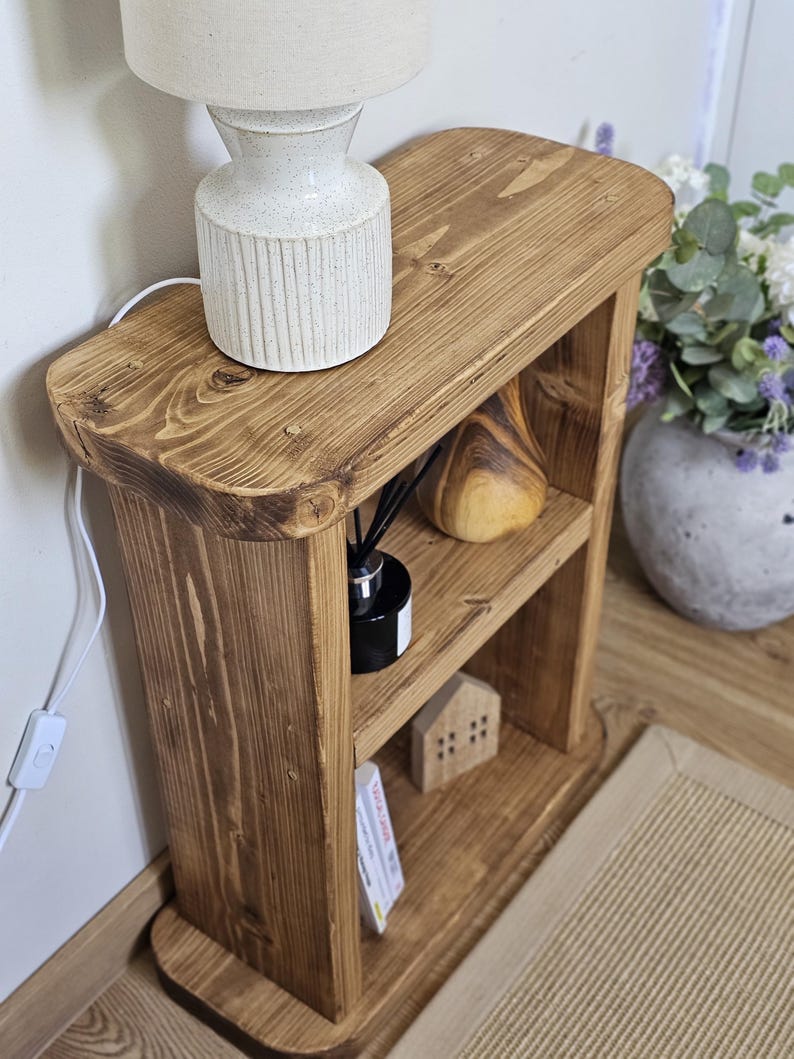 May include: A small, rustic wooden side table with two shelves. A white lamp with a beige lampshade sits on top. The shelves hold a wooden sculpture, a black diffuser, a miniature house, and a small stack of cards. The table is made of light brown wood.