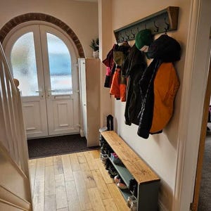 May include: A wooden coat rack with hooks and a shelf, painted in a light green color. The rack is mounted on a wall and has several coats and hats hanging from it. There is a wooden bench with a shelf underneath, also painted in a light green color. The bench is located in a hallway with a wooden floor and a white door.