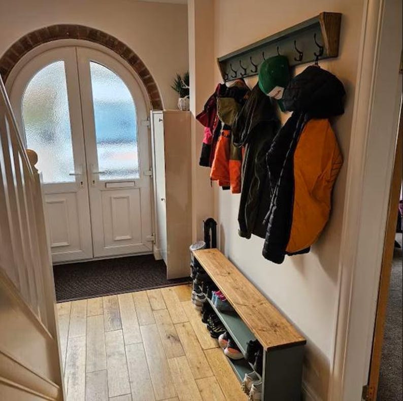 May include: A wooden coat rack with hooks and a shelf, painted in a light green color. The rack is mounted on a wall and has several coats and hats hanging from it. There is a wooden bench with a shelf underneath, also painted in a light green color. The bench is located in a hallway with a wooden floor and a white door.