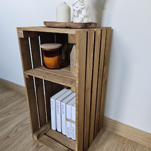 May include: A wooden crate shelf with two shelves. The top shelf has a small white ceramic figurine and a brown candle. The bottom shelf has a stack of books with the words "Home Sweet Home" printed on the spines.
