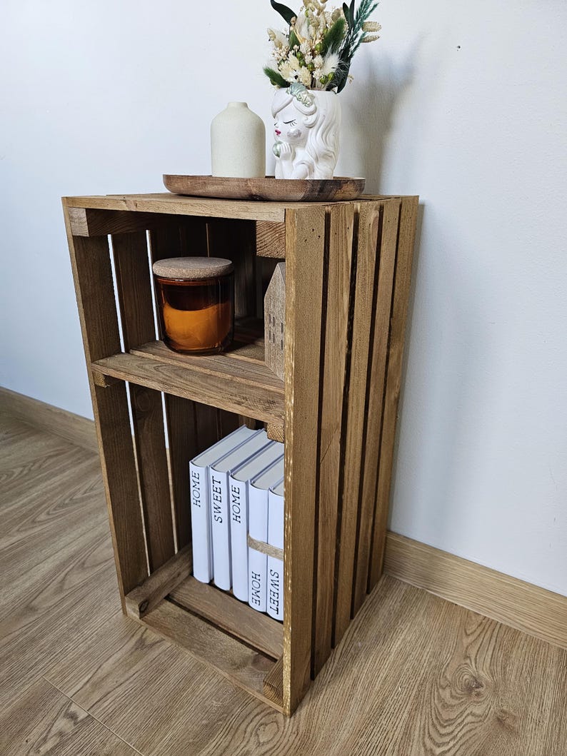 May include: A wooden crate shelf with two shelves. The top shelf has a small white ceramic figurine and a brown candle. The bottom shelf has a stack of books with the words "Home Sweet Home" printed on the spines.