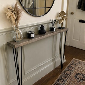 May include: A wooden console table with black hairpin legs. The table displays a vase of dried flowers, a black candle, a black decorative object, a reed diffuser, and a gold-coloured metal sculpture. A round mirror is mounted on the wall above. The table's length is approximately 120 cm.