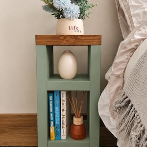 May include: A sage green and brown side table with a vase of flowers and books. The table has two shelves and a wooden top. The vase is cream-colored with the words "Life Life" on it. The books are blue and white.