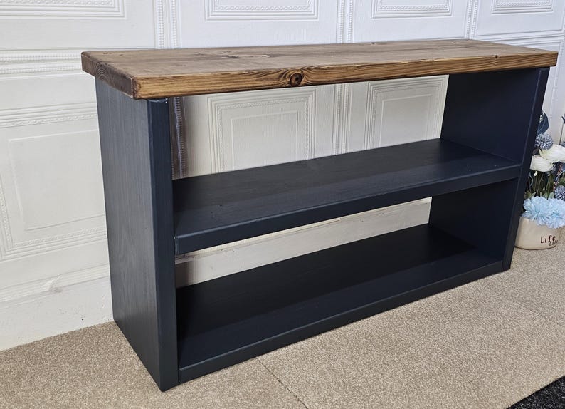 May include: A black wooden shelf unit with two shelves and a rustic wood top. The shelf unit is standing on a beige carpet.