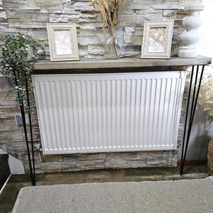 May include: A white radiator with a wooden console table with black metal legs. The table has two framed pictures and a vase with dried flowers. The console table is in front of a stone wall.