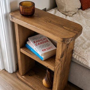 May include: A rustic, wooden side table with two shelves. The top shelf holds a small, amber-colored glass bowl. The middle shelf displays two books, one titled "RADICAL CANDOR". A decorative wooden pear sits on the bottom shelf. The table is made of light brown wood.