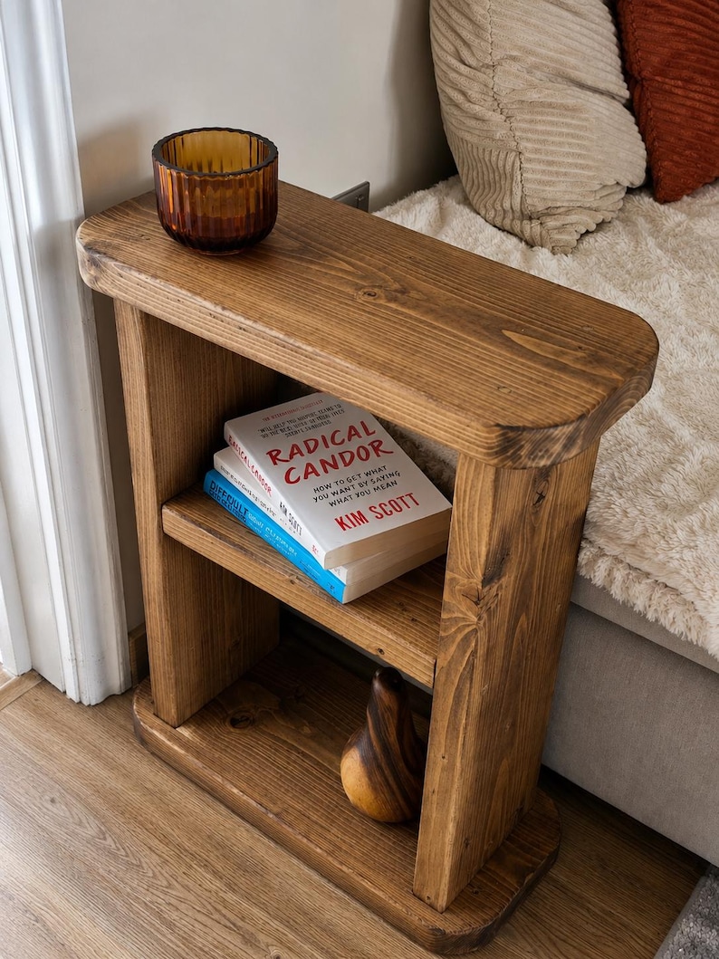 May include: A rustic, wooden side table with two shelves. The top shelf holds a small, amber-colored glass bowl. The middle shelf displays two books, one titled "RADICAL CANDOR". A decorative wooden pear sits on the bottom shelf. The table is made of light brown wood.