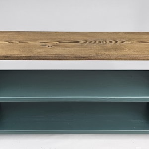 May include: A two-tiered wooden shelf unit with a natural wood top and teal blue painted shelves. The shelf unit is freestanding and has a simple, modern design.
