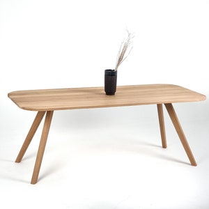 Peut inclure: Une table rectangulaire en bois clair aux coins arrondis avec quatre pieds inclinés. Un vase cylindrique brun foncé est posé au centre du plateau, contenant une tige de plante séchée fine et claire avec des branches délicates.