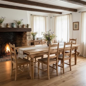 Puede incluir: Una mesa de comedor de madera con cuatro sillas a juego, un camino de mesa de color claro y un centro de mesa floral. El conjunto está situado en una habitación con chimenea de piedra, vigas de madera oscura y cortinas blancas.