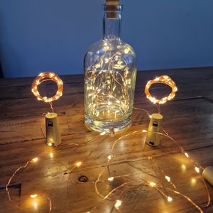 May include: Copper wire fairy lights with warm white LED bulbs wrapped around a glass bottle and two cork stoppers. The lights are strung across a wooden table.