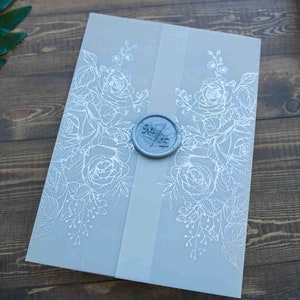 May include: A light gray invitation with white floral designs and a light gray ribbon. A circular, blue-gray wax seal with initials is centered on the invitation. The invitation is on a wooden surface.
