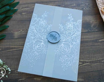 Puede incluir: Una invitación gris claro con diseños florales blancos y una cinta gris claro. Un sello de cera circular azul-gris con iniciales está centrado. La invitación está sobre una superficie de madera.