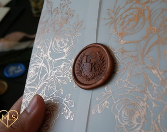 Puede incluir: Una invitación de boda azul claro con un diseño floral de rosas en lámina de cobre. La invitación tiene un sello de cera con las iniciales "L & K" en color cobre.