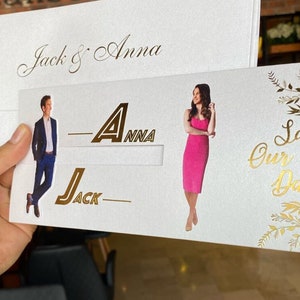 May include: White save the date card with gold lettering. The card features a photo of a couple with the text "Jack & Anna" and "Save Our Date".