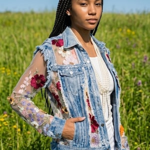 May include: A light blue denim jacket with floral embroidery and sheer sleeves. The jacket features a classic collar, front pockets, and a distressed hem. The embroidery includes red, pink, and purple flowers.