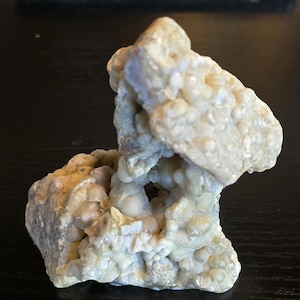 May include: A light-colored, textured rock formation with a bubbly, almost spherical surface. The rock has a rough, uneven texture with a hollowed-out section in the center, creating a cave-like appearance. The overall color is a mix of off-white and pale beige.
