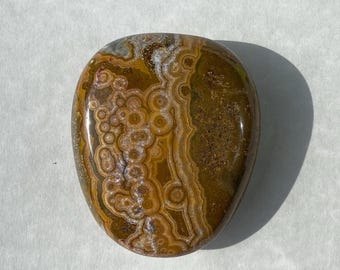 Può includere: Pietra ovale lucidata con un motivo unico di cerchi concentrici e fasce stratificate nei toni del marrone, beige e bianco. La pietra ha una finitura lucida e una consistenza liscia, ideale per la decorazione o il collezionismo.
