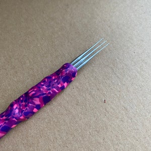 Puede incluir: Un primer plano de una herramienta con tres agujas de metal con un patrón morado y rosa. La herramienta se utiliza para manualidades y creación.