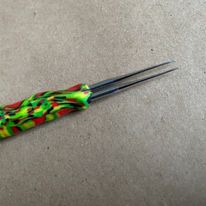 Puede incluir: Un gancho de ganchillo de doble punta con un mango estampado en verde, rojo y negro.