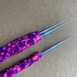 May include: Two crochet hooks with pink and purple speckled handles. The metal tips are silver and pointed. The handles are made of a polymer clay material. The hooks are lying on a brown surface.