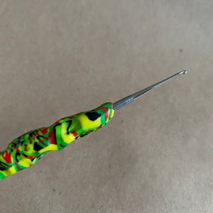 Puede incluir: Un primer plano de un gancho de ganchillo con un mango colorido y estampado. El gancho es de metal y tiene una punta afilada.