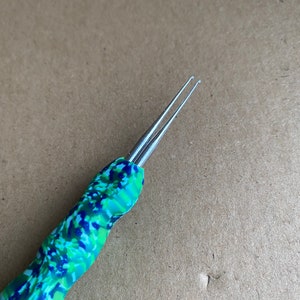 Puede incluir: Un par de agujas de crochet de doble punta con un mango de patrón de camuflaje verde, azul y blanco.