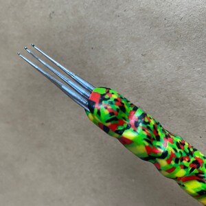 Puede incluir: Primer plano de un gancho de ganchillo multicolor con tres agujas. El gancho es verde, amarillo, rojo y negro.