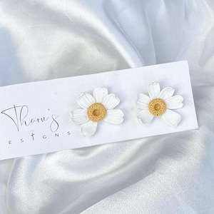 Puede incluir: Un par de pendientes de margaritas blancas con centros amarillos, presentados en una tarjeta blanca con el texto "Thanu's Designs". Los pendientes se colocan sobre un fondo blanco y sedoso, creando una presentación limpia y elegante. Un accesorio floral.