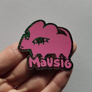 Könnte beinhalten: Ein rosa Anstecknadel mit einem glitzernden grün-schwarzen Rand. Die Anstecknadel zeigt eine stilisierte Elefantenform mit einem Augendesign und dem Wort "Mausig" in einer dekorativen Schriftart.