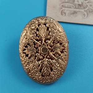 Può includere: Spilla ovale color oro antico con un intricato design traforato a foglie e volute. La spilla è posta su uno sfondo blu, mettendo in risalto la sua dettagliata lavorazione artigianale e l'estetica vintage. Una fotografia d'epoca è sullo sfondo.