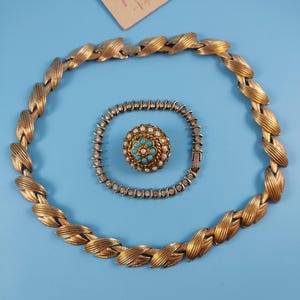 Könnte beinhalten: Ein goldfarbenes Halsketten-, Armband- und Broschen-Set auf blauem Hintergrund. Die Halskette und das Armband weisen ein sich wiederholendes Blattdesign auf. Die Brosche hat ein Blumendesign mit türkisen und Perlenakzenten. Ein Vintage-Schmuckset.