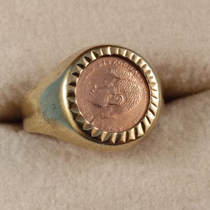 Könnte beinhalten: Ein goldfarbener Ring mit einer kreisförmigen Fassung und gezacktem Rand. In der Mitte des Rings befindet sich eine kupferfarbene Münzeinlage, die das Profil von John Fitzgerald Kennedy zeigt, wobei der Text John Fitzgerald Kennedy sichtbar ist.