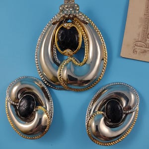 Op de afbeelding: Een sieradenset bestaande uit een grote druppelvormige broche en twee bijpassende ovale clip-oorbellen. Elk stuk heeft een gepolijst zilverkleurig metalen frame, goudkleurige accenten, heldere strass-steentjes en een centrale zwarte cabochon.