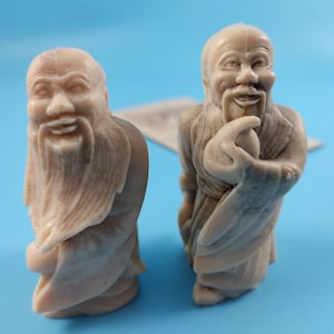 Pode incluir: Duas estátuas em miniatura bege de figuras barbudas sorrindo em trajes. As figuras têm traços faciais detalhados e barbas longas. Elas estão em um fundo azul claro. As estátuas parecem ser feitas de um material semelhante a pedra.