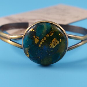 Op de afbeelding: Een armband met een cirkelvormig middendeel met een donkerblauw, groenblauw en groen gemarmerd patroon, geaccentueerd door ingebedde bladgoudvlokken. De armband heeft een antieke messing afwerking en een gespleten bandontwerp.