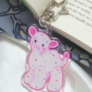 May include: A clear acrylic keychain featuring a white lamb illustration with pink outlines and multicolored polka dots. The lamb has a stitched appearance and is attached to a silver-colored star-shaped metal clasp. The design is flat and glossy.