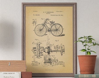 May include: A vintage style print of a bicycle patent drawing with the title "A.T. DeForest, Bicycle, No. 614,455, Patented Nov. 22, 1898" hanging on a wall above a wooden table with a wooden box, a potted plant, and a stack of books.