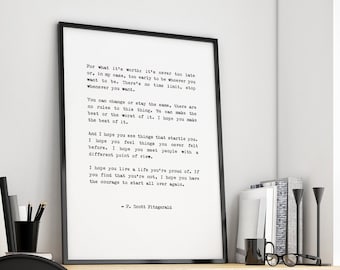 May include: A black and white framed print with a quote by F. Scott Fitzgerald. The quote reads: "You can't repeat the past.  It's gone, it's over.  It's finished.  Why do you keep hanging on to it?" The print is hanging on a white wall above a wooden desk with a black chair, a potted plant, and a pair of glasses.