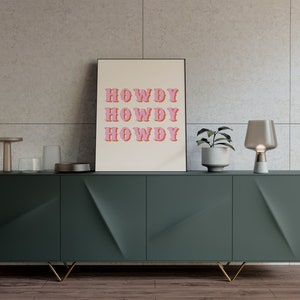 May include: A green cabinet with gold legs and three drawers. A framed print with the text "HOWDY" repeated three times in pink letters is on top of the cabinet. A small plant in a white pot and a gray glass lamp are also on top of the cabinet.