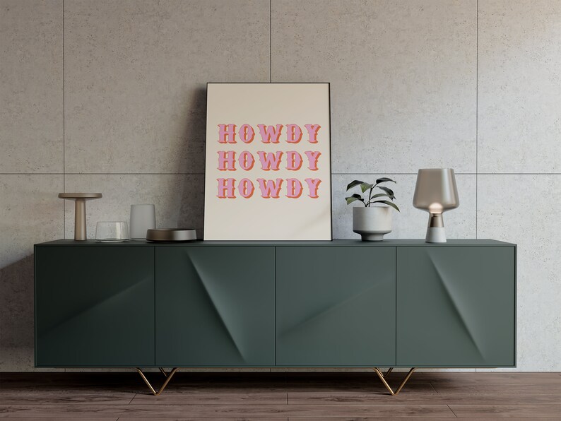 May include: A green cabinet with gold legs and three drawers. A framed print with the text "HOWDY" repeated three times in pink letters is on top of the cabinet. A small plant in a white pot and a gray glass lamp are also on top of the cabinet.